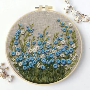 May include: A circular embroidery hoop containing a piece of beige linen fabric. The fabric features hand-stitched embroidery of a dense meadow of small blue and white flowers with green stems and leaves, arranged in a naturalistic cluster.