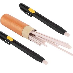 May include: An art supply set featuring a cylindrical container with a brown exterior and a beige interior, filled with numerous light-colored eraser sticks. Two black erasers with yellow buttons are positioned on either side.