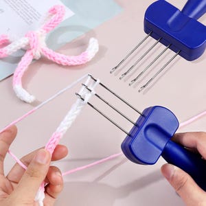 May include: A blue plastic knitting tool with four metal prongs featuring small hooks at the ends. The tool is used to create a white and pink braided yarn cord. A finished pink and white yarn bow is visible in the upper left corner of the frame.