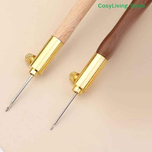 May include: Two embroidery tools with wooden handles and gold-colored metal components. One tool has a light wood handle, and the other has a dark wood handle. The tools have a needle extending from the metal component.