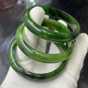 Könnte beinhalten: Drei grüne Jade-Armreifen in verschiedenen Grüntönen sind übereinander gestapelt. Die polierten Steinarmbänder haben eine glatte, glänzende Oberfläche. Die Armreifen werden auf einer weißen Oberfläche präsentiert.