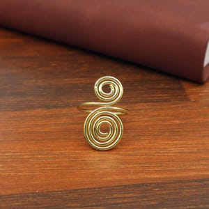 May include: A gold-coloured metal ring featuring two connected spiral designs. The ring is constructed from a single continuous wire that forms a smaller spiral at the top and a larger spiral at the bottom, creating an open, adjustable band structure.