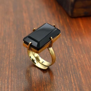 May include: A gold-toned ring featuring a large, rectangular, faceted black stone set in a four-prong setting. The band is smooth and metallic, showcasing a polished finish that complements the dark, reflective surface of the central gemstone.