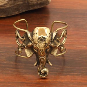 May include: A gold-toned metal cuff bracelet featuring a central, detailed elephant head design. The elephant has a prominent trunk, large ears, and decorative engravings on its forehead. The band is open-ended with thin, curved metal wires.