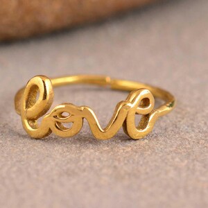 Könnte beinhalten: Ein goldfarbener Ring mit dem Wort love in durchgehender Schreibschrift. Das Metallband ist dünn und kreisförmig, wobei die Buchstaben den Mittelpunkt des Designs bilden. Die Oberfläche des Rings hat ein glattes, poliertes Finish.