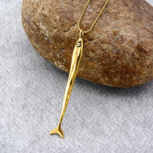 May include: A gold-colored fish pendant necklace. The pendant is a stylized fish with a textured surface and a small eye detail. The necklace chain is also gold-colored and delicate. The pendant is approximately 3 inches long.