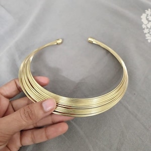 May include: A gold-colored collar necklace with multiple stacked wire strands. The necklace is held by a hand against a gray background. The necklace has a simple, elegant design.