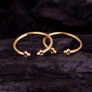 May include: Two gold-toned open hoop rings, each featuring a small cluster of three tiny spheres at both ends of the band. The rings have a smooth, polished finish and are positioned side by side, showcasing their circular shape and open design.