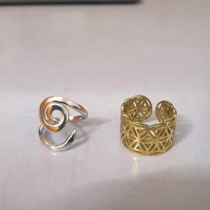 May include: A silver-colored ring featuring a spiral design and a gold-colored ring with a wide band and a geometric, repeating floral-like cutout pattern. Both rings are open-ended, allowing for adjustable sizing on the finger.