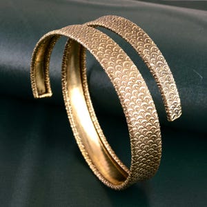 May include: Two gold-colored bangles with an intricate, repeating pattern. The open-ended design allows for easy wear. The bangles are displayed against a dark green background, highlighting their metallic sheen.