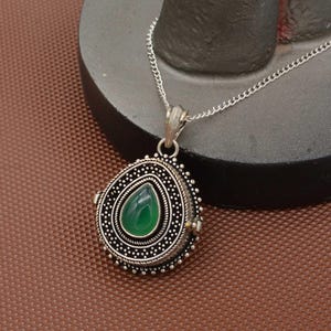 May include: Silver-toned pendant necklace with a teardrop-shaped green stone. The pendant has an ornate, detailed design with a beaded border and a delicate chain. The necklace is displayed on a brown surface.
