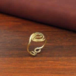 Puede incluir: Un anillo de metal en tono dorado con un diseño de banda abierta y ajustable. La parte superior del anillo muestra un patrón de alambre continuo y en espiral que forma una figura circular. El metal tiene un acabado liso y pulido que refleja la luz.