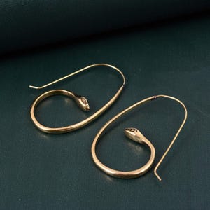 May include: A pair of gold-colored snake-shaped earrings. The earrings feature a coiled snake design with a small head and a curved body. The earrings are made of a shiny metal and are displayed on a dark green surface.