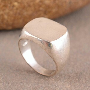 Könnte beinhalten: Ein silberfarbener Siegelring mit einer glatten, flachen, quadratischen Oberfläche mit abgerundeten Ecken. Die Ringschiene ist massiv und verjüngt sich leicht nach unten, wobei das gesamte Schmuckstück ein poliertes Metallfinish aufweist.