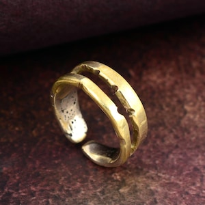 May include: A gold-colored adjustable ring with a double-band design. The ring has a textured surface and an open, adjustable design. The ring is made of metal and is suitable for everyday wear. The ring is shown against a dark brown background.