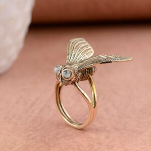 Può includere: Un anello color oro a forma di mosca. La mosca ha ali dettagliate e due pietre rotonde e trasparenti per gli occhi. La fascia dell'anello ha un design semplice e liscio.