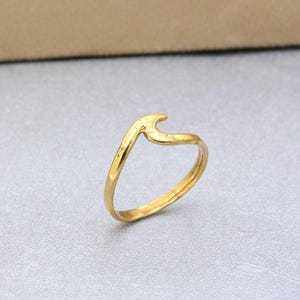 Può includere: Un anello color oro caratterizzato da un design a onda singola, liscia e stilizzata sulla parte superiore. La fascia è sottile e circolare, con una finitura metallica lucida che riflette la luce. L'elemento a onda si curva verso l'alto.