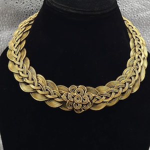 Op de afbeelding: Een goudkleurige metalen ketting met een doorlopend gevlochten draadontwerp dat een cirkelvormige kraag vormt. Het midden van het sieraad is versierd met een cluster van gewikkelde draadspiralen, wat zorgt voor een getextureerd en gelaagd uiterlijk.