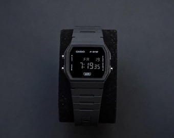 Puede incluir: Un reloj digital negro Casio F-91W con una esfera rectangular que muestra la hora 7:19:35 y el día, viernes 19. El reloj está sobre un soporte negro sobre un fondo oscuro. El reloj tiene una correa negra.