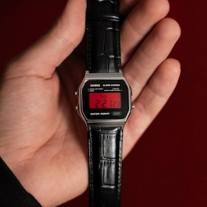 May include: A digital wristwatch with a silver-tone rectangular case and a black faux-leather strap featuring a crocodile-embossed texture. The watch face displays a red digital screen with the text CASIO, ALARM CHRONO, and WATER RESIST.
