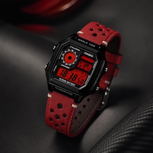 May include: A black digital watch with a red display and a red perforated leather strap. The watch face displays the time, date, and other functions. The watch is branded with the name "CASIO".