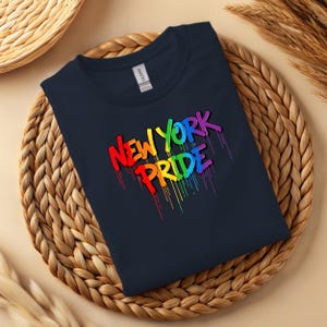 May include: Navy blue t-shirt with the words "New York Pride" in a rainbow-colored, dripping paint design. The shirt is displayed on a woven, circular placemat.