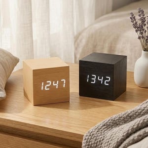 May include: Two cube-shaped digital clocks. One is light wood, displaying "1247" in white. The other is black, showing "1342". Both clocks sit on a wooden surface, with a pillow, vase with flowers, and a blanket in the background.