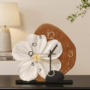 May include: A decorative clock featuring a white flower design with a wooden background. The clock has black hands and a black base. The numbers are in a gold color.