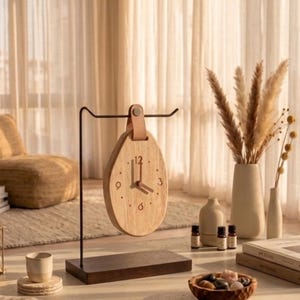 May include: A wooden teardrop-shaped clock with a light brown leather strap hangs from a black metal stand. The clock face has black numerals and hands. The clock sits on a dark brown wooden base. The scene is a neutral-toned interior with decorative elements.