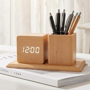 Peut inclure: Un organiseur de bureau en bois avec une horloge numérique affichant 12h00 et un porte-stylos. L'horloge et le porte-stylos sont reliés sur une base en bois. Le porte-stylos contient plusieurs stylos. L'organisateur est en bois clair.