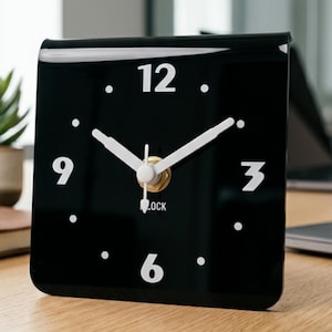 May include: A square black clock with white numerals and hands. The clock face has the numbers 3, 6, 9, and 12, with dots marking the other hours. The word "LOCK" is printed below the hands.