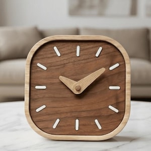 Peut inclure: Une horloge carrée en bois aux coins arrondis. Le cadran est en bois brun foncé avec des marqueurs blancs et des aiguilles en bois brun clair. L'horloge est posée sur une surface en marbre blanc.