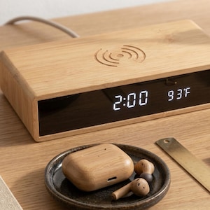 May include: A wooden wireless charging alarm clock displaying 2:00 and 93°F. The clock has a wireless charging symbol on top. A small dish holds wooden earbuds and a charging case. A brass ruler is also present.