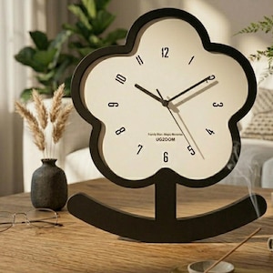 May include: A black and white flower-shaped desk clock with a rocking base. The clock face is off-white with black numerals and hands. The text "Family Von-Mighty Moments UG2DOM" is printed on the clock face. The clock is on a wooden surface.