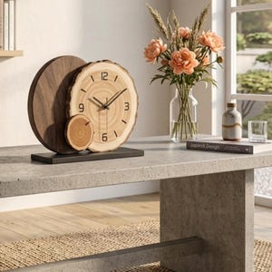 Puede incluir: Un reloj de mesa con un panel circular grande de madera oscura y una rodaja de madera clara más pequeña con bordes de corteza natural. La esfera muestra números negros en el 3, 6, 9 y 12, con manecillas negras sobre una base rectangular.