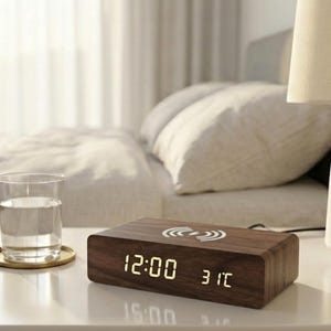 May include: A rectangular wooden digital clock displaying 12:00 and 31°C. The clock has a wireless charging symbol on top. A glass of water sits on a gold coaster to the left of the clock. The background includes a bed and a lamp.