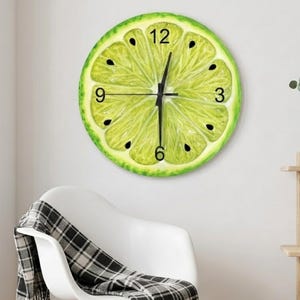 Pode incluir: Um relógio de parede redondo com um design de fatia de limão. O mostrador do relógio apresenta um segmento de limão verde e amarelo com sementes pretas e ponteiros pretos. O relógio está montado numa parede branca.