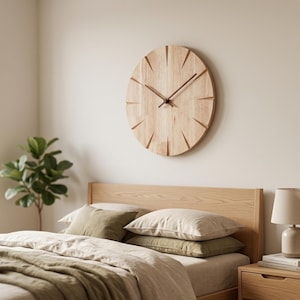 Op de afbeelding: Een ronde houten wandklok met een lichte houtnerf afwerking hangt boven een bed. De klok heeft een minimalistisch ontwerp met uitgesneden uurmarkeringen en donkerbruine wijzers. Het bed heeft een houten hoofdbord, beige beddengoed en groene kussens.