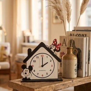 May include: A house-shaped clock featuring Mickey Mouse and Minnie Mouse. The clock face is white with black numbers and hands. The clock is on a wooden table, with a vase and books in the background.