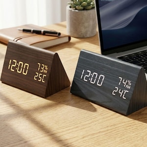 Puede incluir: Dos relojes digitales triangulares de madera. Uno es marrón oscuro con dígitos ámbar, mostrando la hora, la humedad y la temperatura. El otro es negro con dígitos blancos. Ambos están sobre una superficie de madera.