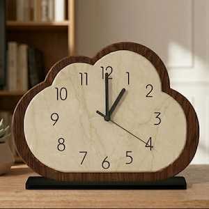 Peut inclure: Horloge de bureau en forme de nuage avec un cadran beige clair et un cadre en bois marron foncé. L'horloge comporte des chiffres et des aiguilles noirs et repose sur une base noire. L'horloge mesure environ 20 cm de large et 18 cm de haut.
