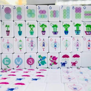 May include: A set of white Mahjong tiles featuring colorful illustrations including concentric circles, potted bamboo plants, lanterns with Chinese characters, compass directions, flowers, and monkeys holding umbrellas with the text JOKER, BAM, and CRAK.