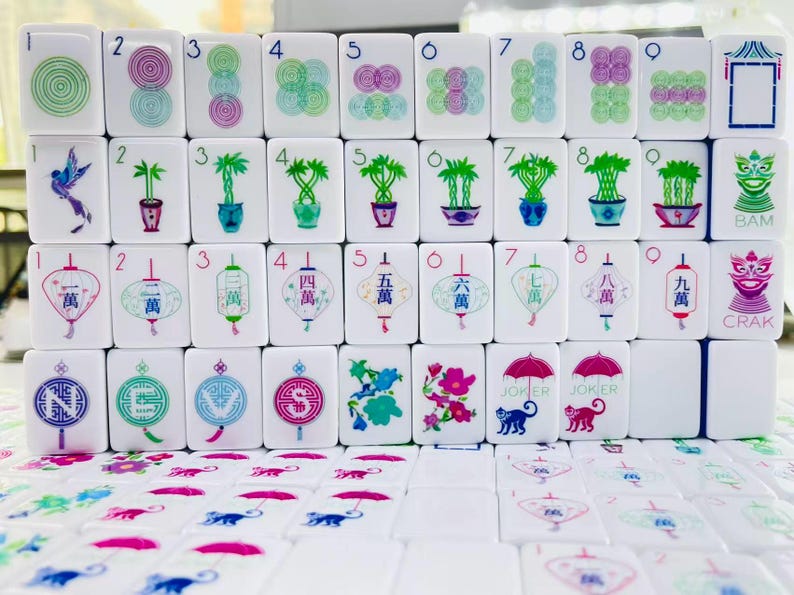 May include: A set of white Mahjong tiles featuring colorful illustrations including concentric circles, potted bamboo plants, lanterns with Chinese characters, compass directions, flowers, and monkeys holding umbrellas with the text JOKER, BAM, and CRAK.