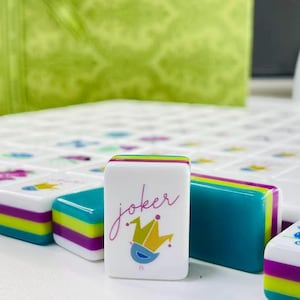 Puede incluir: Una ficha de mahjong rectangular blanca con la palabra joker en letra cursiva sobre un gr&aacute;fico de corona estilizada en amarillo y azul. La ficha tiene bordes laterales en capas de color verde azulado, verde lima y morado.