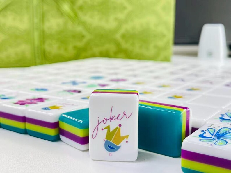 Puede incluir: Una ficha de mahjong rectangular blanca con la palabra joker en letra cursiva sobre un gr&aacute;fico de corona estilizada en amarillo y azul. La ficha tiene bordes laterales en capas de color verde azulado, verde lima y morado.