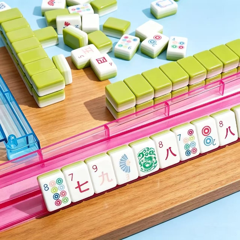 May include: A Mahjong set featuring rectangular tiles with lime green backs and white faces displaying various colorful symbols, numbers, and Chinese characters. The tiles are arranged in rows on a wooden surface, supported by translucent pink and blue plastic racks.