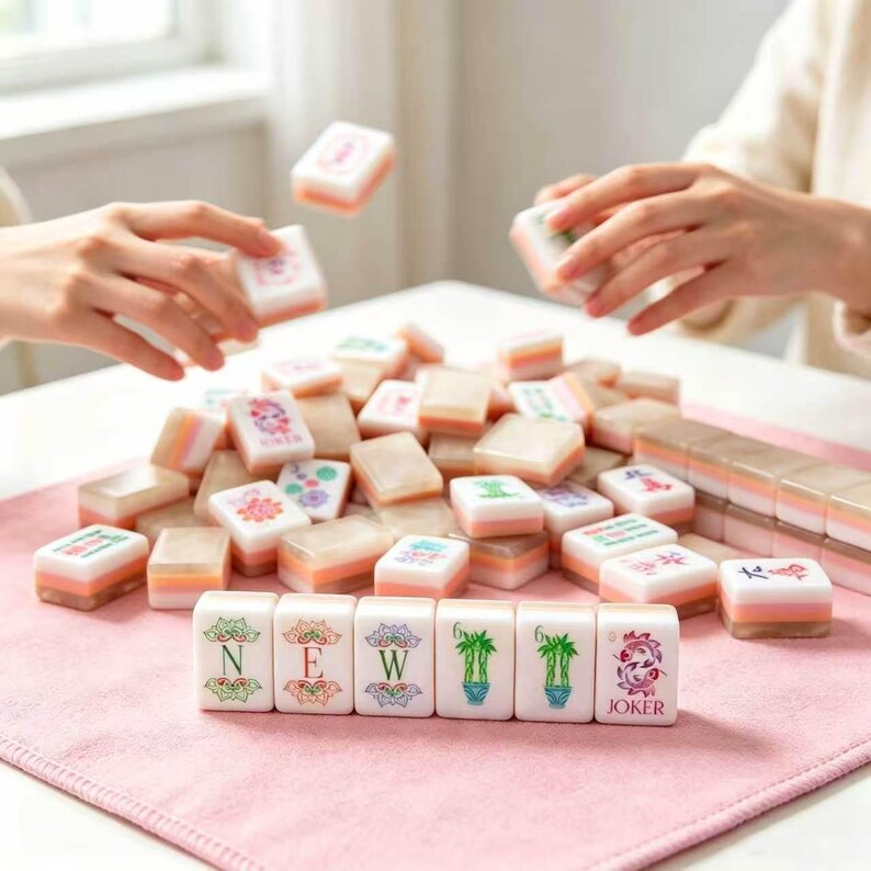 May include: A collection of rectangular Mahjong tiles with white faces and pinkish-orange backs. The front row displays tiles with green floral designs, the letters N, E, W, and the word JOKER in pink. Other tiles are scattered across a pink mat.