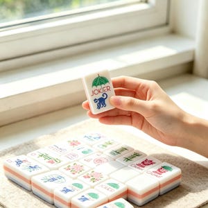 May include: A rectangular white mahjong tile featuring a green umbrella graphic, the word JOKER in red text, and a blue monkey illustration. The tile has a layered base with white, pink, and light blue stripes, matching other tiles arranged nearby.