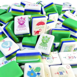 May include: A collection of Mahjong tiles featuring white faces with colorful engraved symbols, including characters, numbers, and a joker, layered over green and blue striped bases. The tiles display various icons like a monkey, umbrella, and floral patterns.