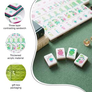 May include: A set of white acrylic mahjong tiles featuring colorful floral and character designs, including tiles labeled Joker, Crak, and Bam. The tiles have a three-layer contrasting sandwich edge and are presented with a green gift box.